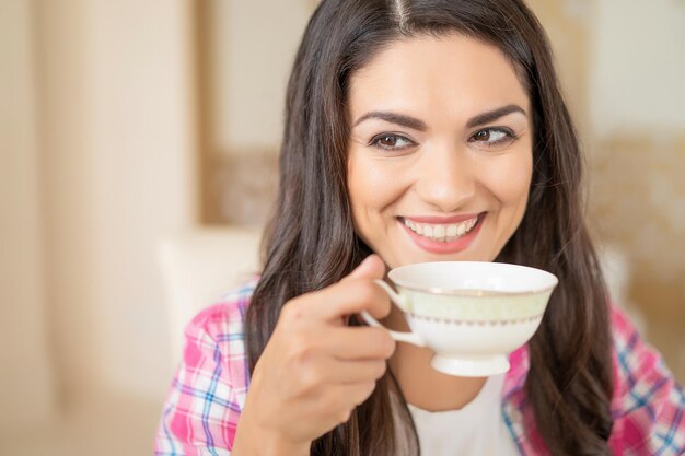 Chica morena sonriente bebe un café de una taza en un restaurante mientras mira de lado y coquetea