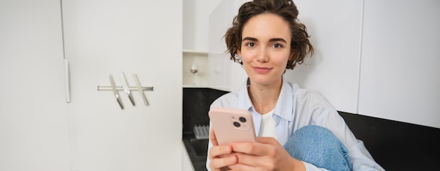 Una chica morena sonriente sentada con un teléfono inteligente en casa ordena comida para llevar desde una aplicación de teléfono móvil usando