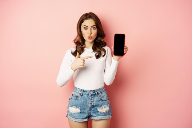 Chica morena sorprendida apuntando a la pantalla de la aplicación de teléfono móvil, mostrando anuncios de teléfonos inteligentes, oferta de compras en línea, de pie asombrada contra el fondo rosa.