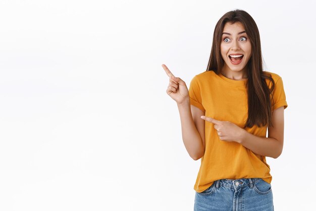 Chica morena sorprendida y asombrada ve a una famosa celebridad señalando y mirando emocionada en la esquina superior izquierda sonriendo con una expresión impresionada y asombrada de pie fascinado de fondo blanco