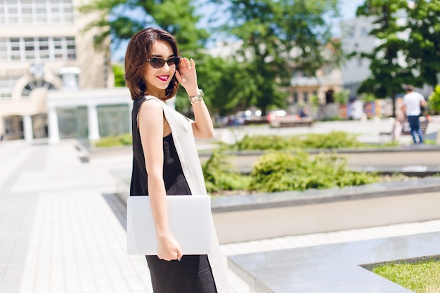 Chica morena vestida de negro y gris está caminando en el parque. Ella sostiene gafas de sol negras en la cara y una computadora portátil en la mano. Ella está sonriendo con labios vinosos.