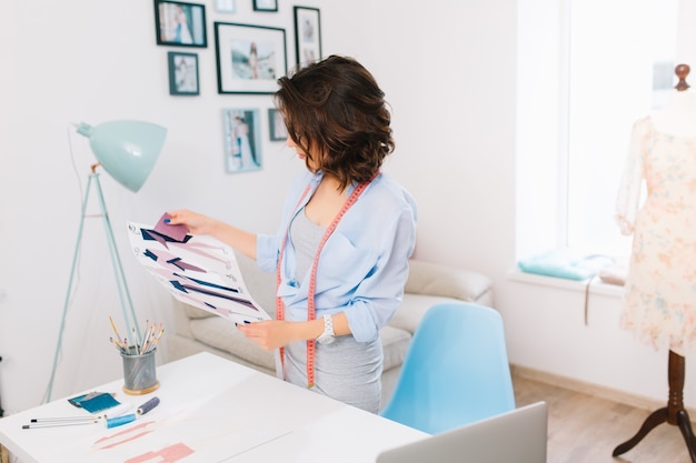 Una chica morena con un vestido gris y una camisa azul está de pie cerca de la mesa en un taller de estudio. Ella está mirando algunas muestras de material y bocetos en sus manos.