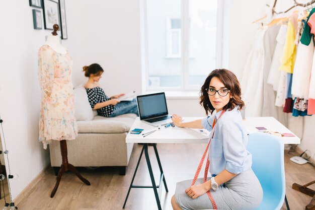 Una chica morena con un vestido gris y una camisa azul está sentada a la mesa en el estudio del taller. Ella sonríe a la cámara. Otra chica está sentada en el sofá en el fondo.