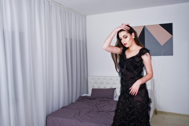 Chica morena en vestido de noche negro contra la habitación blanca con cama