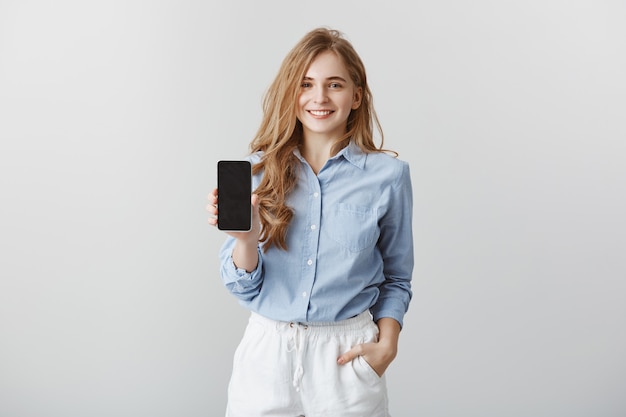 Chica mostrando nuevo teléfono a un colega. Retrato de una encantadora bloguera de moda europea de aspecto amistoso en una blusa azul formal, sosteniendo la mano en el bolsillo mientras muestra el teléfono inteligente sobre una pared gris, publicidad