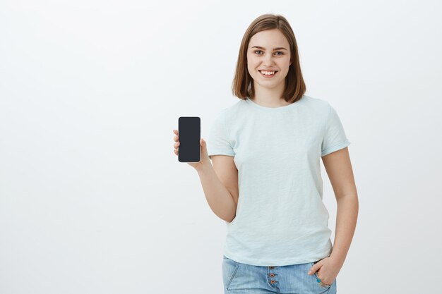 Chica mostrando nuevo teléfono que los padres compraron para el nuevo período escolar. Encantada y complacida joven encantadora mostrando la pantalla del teléfono inteligente hablando de una aplicación genial que encontró en la tienda en línea sobre una pared gris