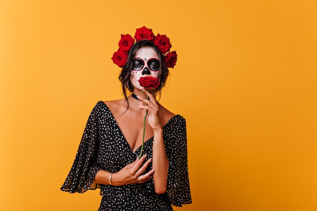 Chica muerta romántica en vestido negro con rosa roja. Retrato interior de mujer zombie lleva corona de flores.