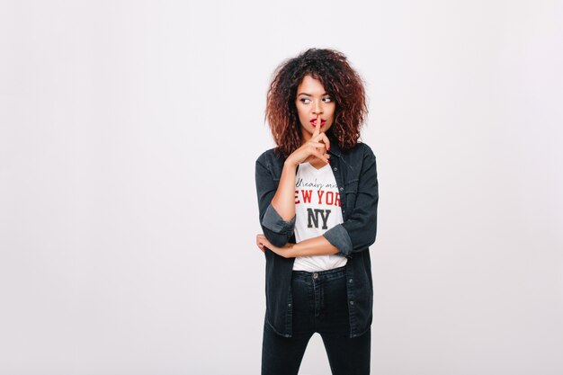 Chica mulata pensativa con cabello castaño rizado mirando a otro lado tocando los labios con el dedo. Retrato interior de elegante modelo femenino con maquillaje brillante en jeans aislado.