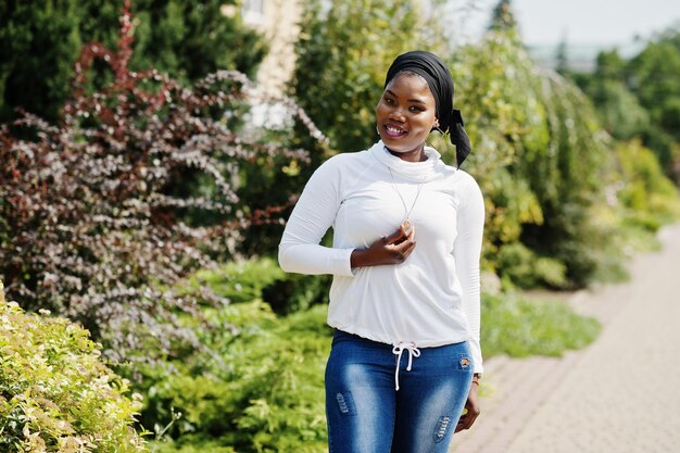 Chica musulmana africana con sudadera blanca hiyab negra y jeans posando al aire libre