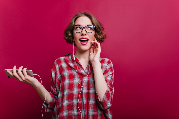 Chica muy divertida con cabello oscuro escuchando música en auriculares blancos y mirando a otro lado. Foto interior de encantadora joven con gafas de moda disfrutando de la canción en la pared clarete.