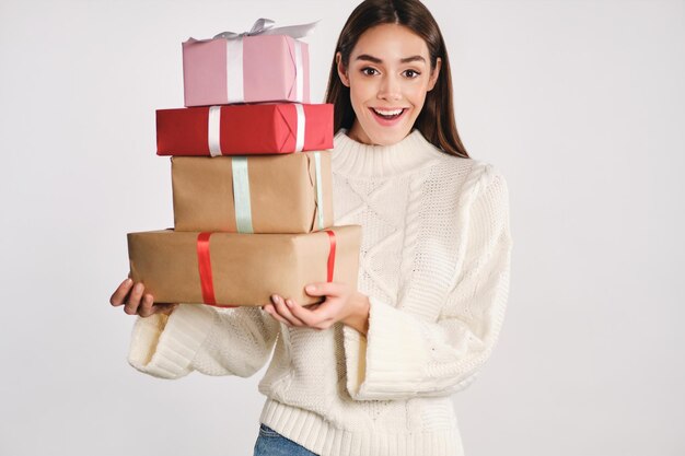 Una chica muy emocionada con un suéter acogedor sosteniendo cajas de regalo mirando alegremente a la cámara sobre fondo blanco