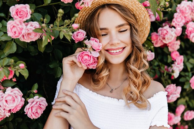 Chica muy europea sonriendo con los ojos cerrados sobre la naturaleza. Hermosa mujer rubia disfrutando de la sesión de fotos con flores.