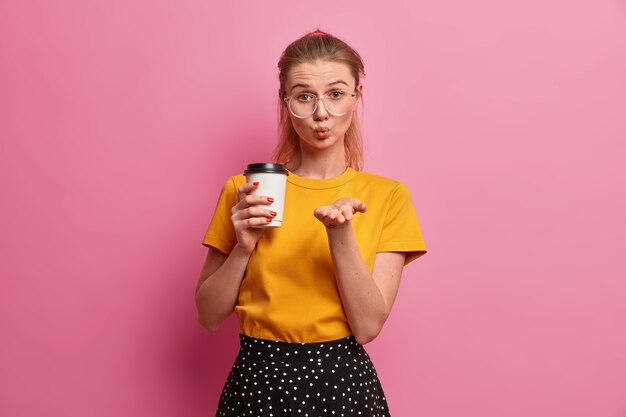 Una chica muy romántica solo envía amor, mantiene los labios redondeados, envía mwah, sostiene café para llevar, se viste de manera informal