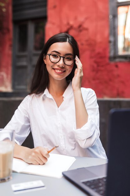 Chica muy sonriente con cabello oscuro en camisa blanca y anteojos mirando felizmente en cámara con computadora portátil y bloc de notas en el acogedor patio de la cafetería