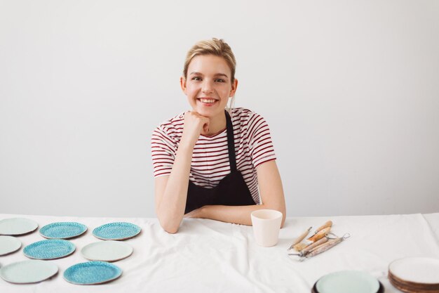 Chica muy sonriente con delantal negro y camiseta a rayas sentada en la mesa con tazas y platos hechos a mano felizmente mirando en cámara al estudio de cerámica