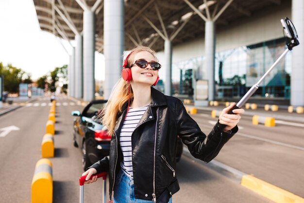 Chica muy sonriente con gafas de sol y auriculares tomando fotos en el celular con un auto descapotable en el fondo