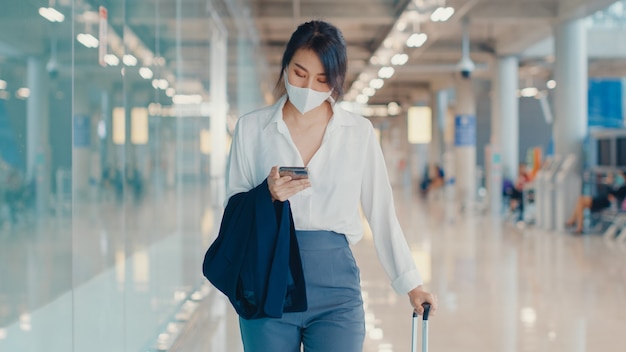 chica de negocios asiática que usa el teléfono inteligente para verificar la tarjeta de embarque caminando con el equipaje a la terminal en el vuelo nacional en el aeropuerto.