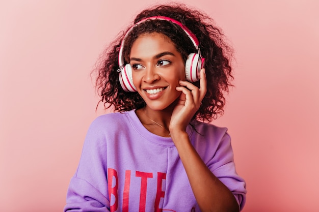 Chica negra alegre escuchando música en colores pastel. Encantadora mujer africana en suéter morado posando en auriculares.