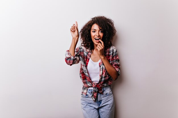 Chica negra alegre con peinado rizado divertido posando en blanco. Señora guapa africana de pie con una sonrisa suave.