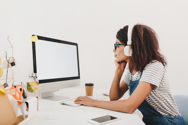 Chica negra delgada en auriculares mirando la pantalla del ordenador con atención