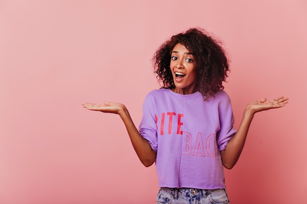 Chica negra de ojos muy oscuros en rosa. Mujer alegre de pelo corto en camisa morada que expresa asombro.