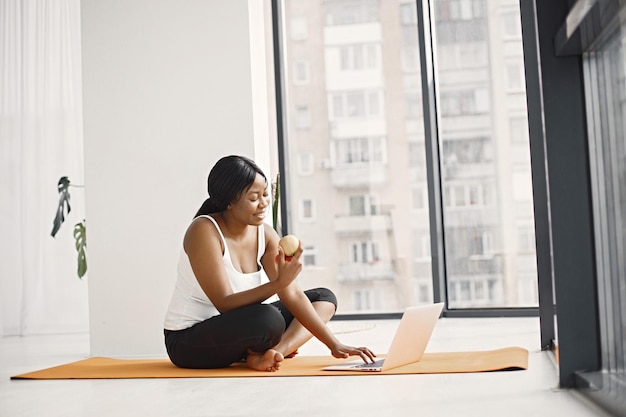 Chica negra sentada en una alfombra de yoga naranja en un estudio con una ventana grande y usando una laptop