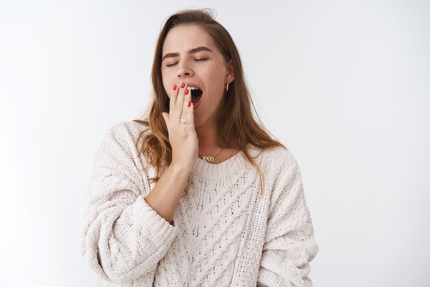 Chica no madrugada. Linda mujer caucásica optimista con suéter acogedor bostezando despreocupadamente ojos cerrados cubriendo la palma de la boca abierta, sintiéndose somnoliento al despertarse después de la siesta, de pie fondo blanco