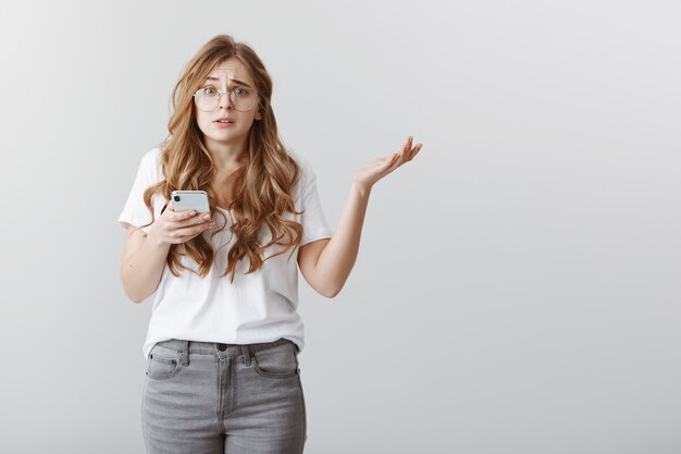 La chica no tiene ni idea de quién envió un mensaje ridículo. Retrato de mujer atractiva confundida preocupada con cabello rubio con gafas, encogiéndose de hombros, extendiendo la palma en gesto inconsciente, sosteniendo el teléfono inteligente, estando nervioso