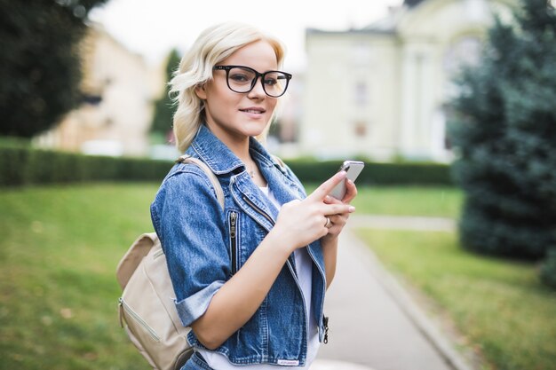 Chica ocupada joven rubia usa el teléfono para desplazarse por la conversación de la red social en la mañana de la plaza otoño de la ciudad