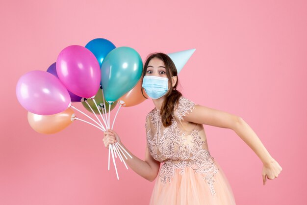 Chica de ojos abiertos con gorra de fiesta corriendo mientras sostiene globos de colores en rosa