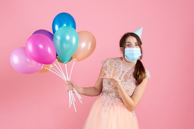 Foto gratuita chica de ojos abiertos con gorro de fiesta sosteniendo globos de colores en rosa