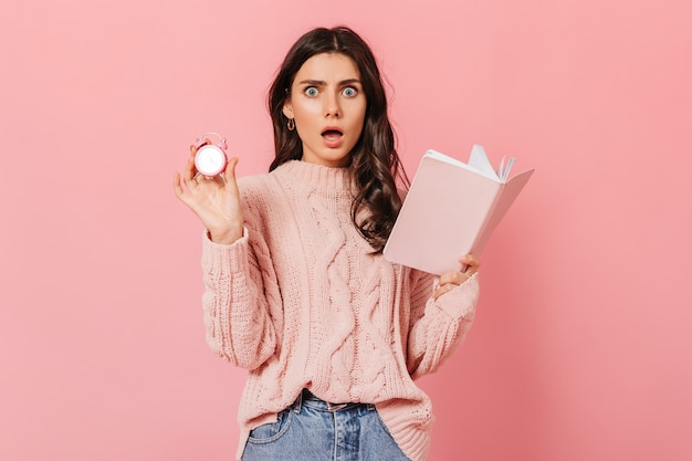 Chica de ojos azules en estado de shock mira a cámara sobre fondo rosa. Dama de suéter elegante posando con despertador y diario.