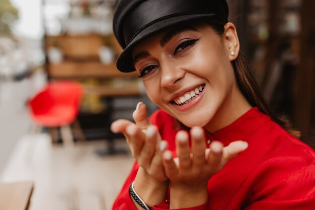 Chica de ojos marrones bellamente pintados con delineador de ojos, destacando los rasgos del rostro. Modelo en blusa roja envía beso al aire