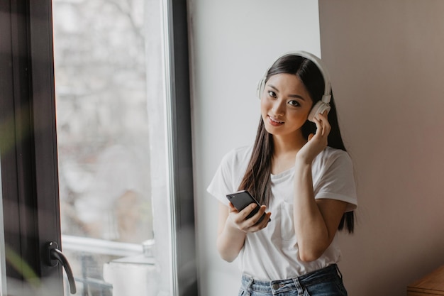 Chica de ojos marrones mira a la cámara y sostiene el teléfono Mujer asiática en la parte superior blanca disfrutando de la canción en los auriculares