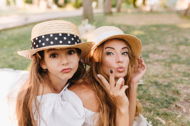 Chica de ojos oscuros con sombrero jugando con mamá disfrutando del fin de semana familiar en el parque verde. Mujer agraciada lleva elegante anillo haciendo muecas y bromeando con hija descansando al aire libre.