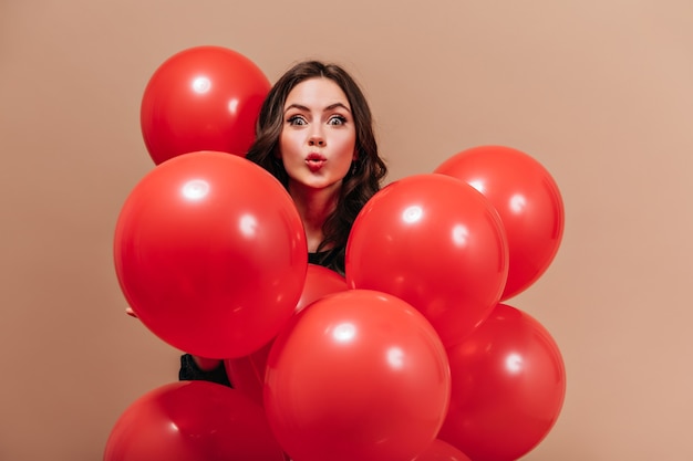 La chica de ojos verdes mira a la cámara con sorpresa, sosteniendo globos rojos sobre fondo beige.