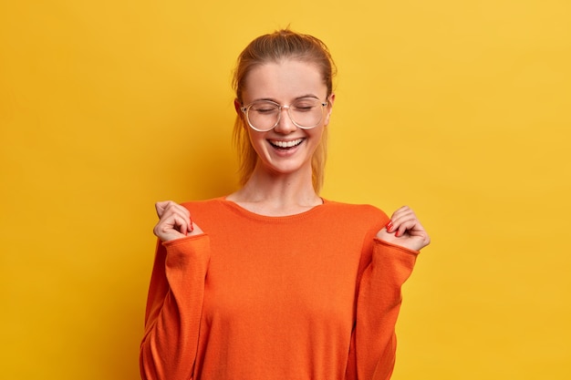 Chica optimista alegre con sonrisa feliz, cierra los ojos, levanta las manos, se ríe por algo positivo, usa gafas ópticas y un jersey naranja,