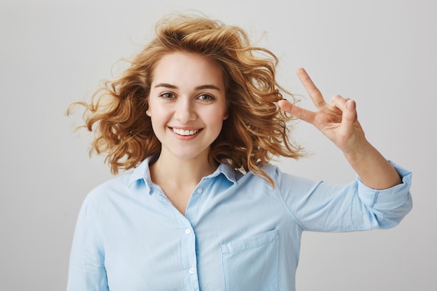 Chica optimista despreocupada con cabello rizado mostrando el signo de la paz y sonriendo