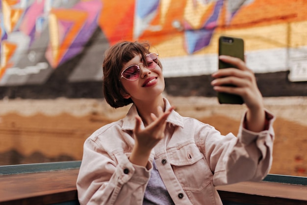 Chica optimista haciendo selfie en la calle Mujer con cabello corto moreno en sunlasses brillantes y chaqueta de mezclilla beige sonriendo afuera