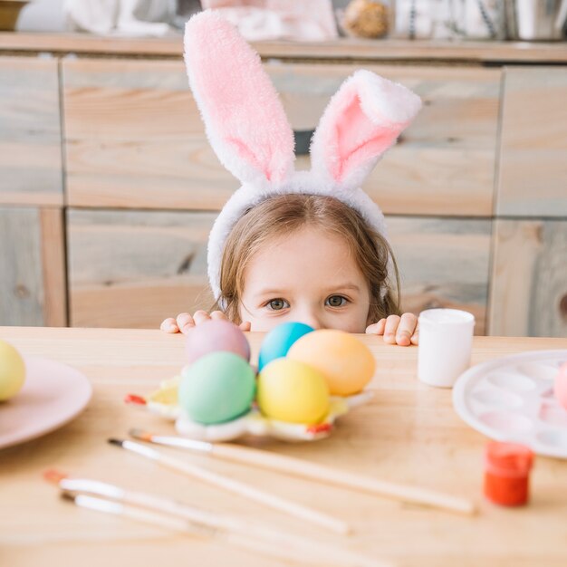 Chica en orejas de conejo escondida detrás de la mesa con huevos de Pascua