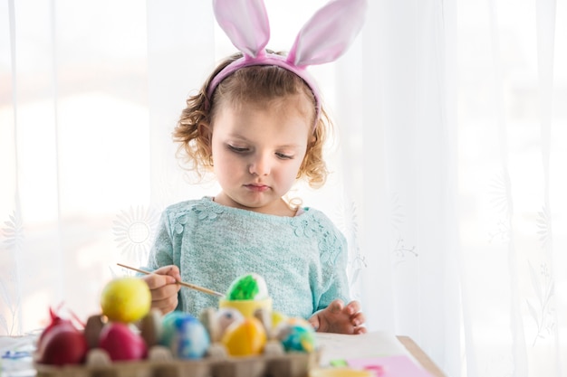 Chica en orejas de conejo pintando huevos de Pascua