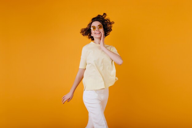 Chica pálida con expresión de la cara de éxtasis disfrutando de la sesión de fotos en traje de verano blanco. Mujer joven complacida en camiseta amarilla sonriendo mientras posa en la pared naranja.