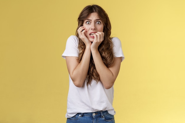 Chica en pánico sintiéndose asustado tímido inseguro morderse las uñas mirar la cámara asustado apretar los dientes temblando miedo tocar la cara nerviosamente sintiéndose inseguro aterrorizado soporte fondo amarillo