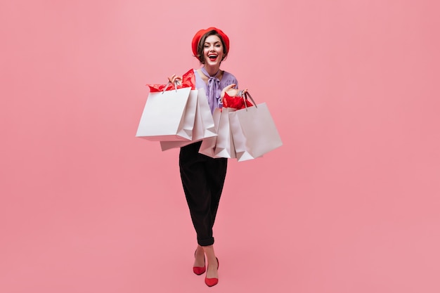 Chica en pantalón negro y boina roja tiene variedad de paquetes y sonríe sobre fondo rosa.