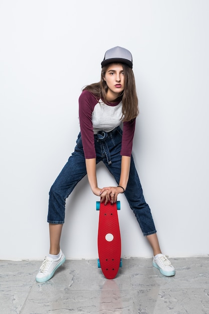 Chica patinadora moderna sosteniendo patineta roja en el piso aislado en la pared blanca