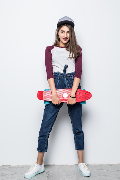 Chica patinadora moderna sosteniendo patineta roja en sus manos aisladas en la pared blanca