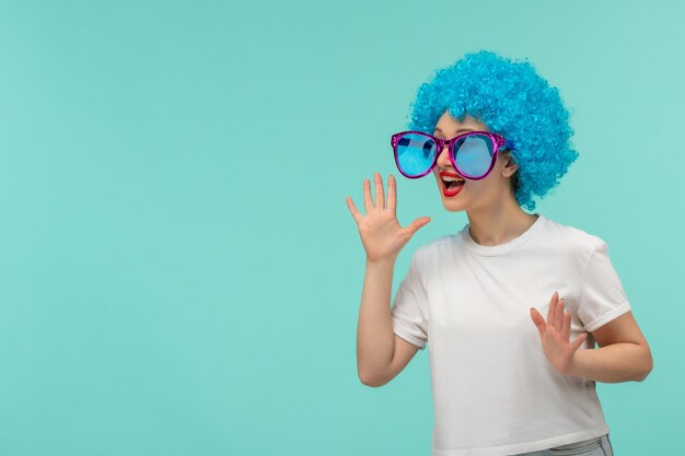 Chica de payaso del día de los inocentes llamando a alguien con las manos en la boca grandes gafas de sol azules traje divertido cabello azul