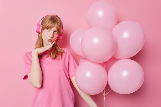 Foto gratuita chica pecosa pelirroja de ensueño aplica parches de belleza debajo de los ojos mantiene los labios doblados escucha música a través de auriculares vestida con una camiseta casual sostiene un montón de globos aislados sobre fondo rosa