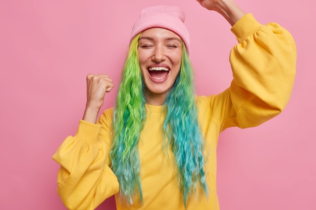 Chica con peinado de moda hace gesto ganador celebra logros exclama de alegría viste sombrero y puente amarillo tiene piercing en la nariz aislado en rosa