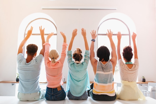 Chica con peinado de moda sentada con las manos en alto junto a sus amigos de la universidad y mirando a la ventana grande. estudiantes felices divirtiéndose en la habitación luminosa después de los exámenes.